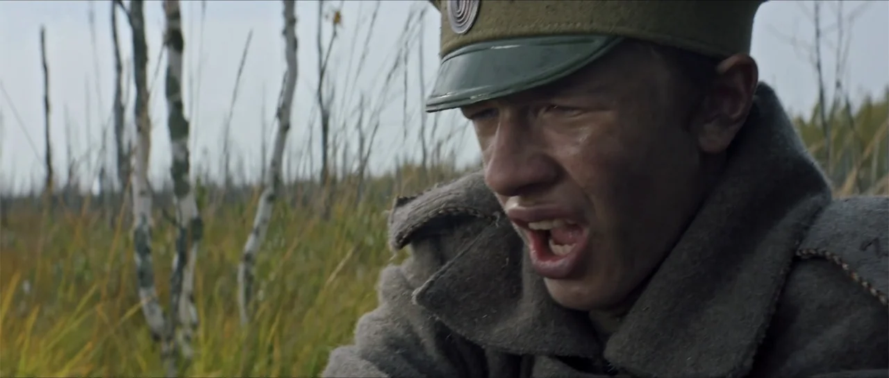 The Rifleman (2019) movie still: closeup — A wetland landscape filled with tall dry grasses and thin birch trees. A young soldier wearing a wool…