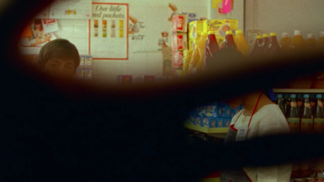 Shirkers (2018) movie still: medium — A small retail shop interior filled with various beverage bottles and packaged food items. The woman stands…