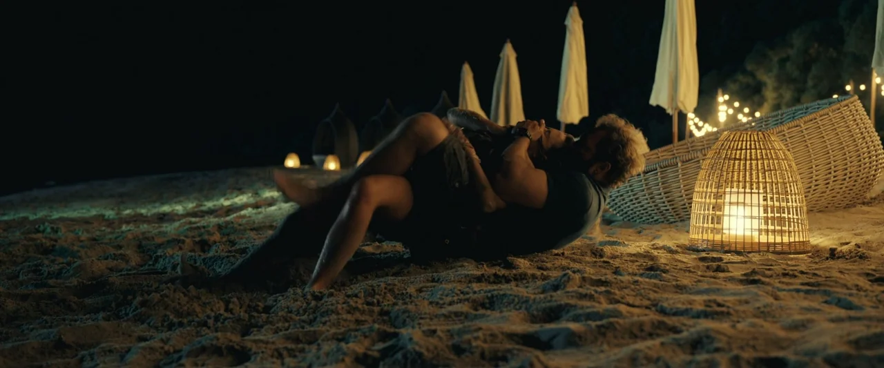 Dangerous Animals (2025) movie still: fullbody — A dark tropical beach at night features woven lamps and umbrellas on the sand. A couple lies in an…