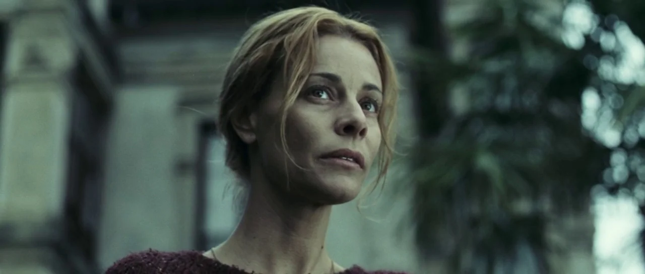 The Orphanage (2007) movie still: closeup — A woman stands before the blurred facade of a large, weathered residential building. She gazes upwards…