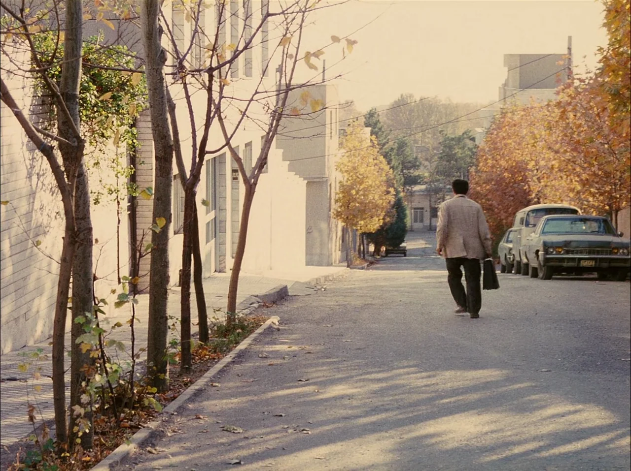 Close-Up (1990) movie still: fullbody — A quiet residential street is lined with young trees and autumn foliage. A man carries a briefcase while…