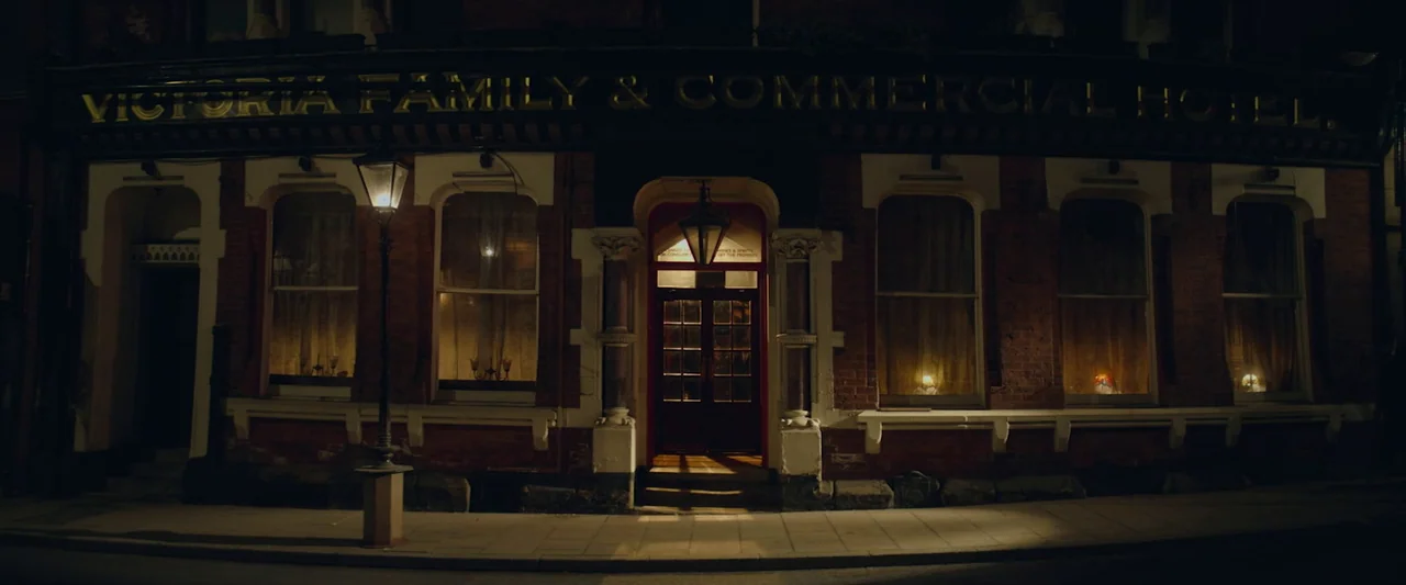 Nandor Fodor and the Talking Mongoose (2023) movie still: establishing — A historic brick building labeled Victoria Family & Commercial Hotel stands…