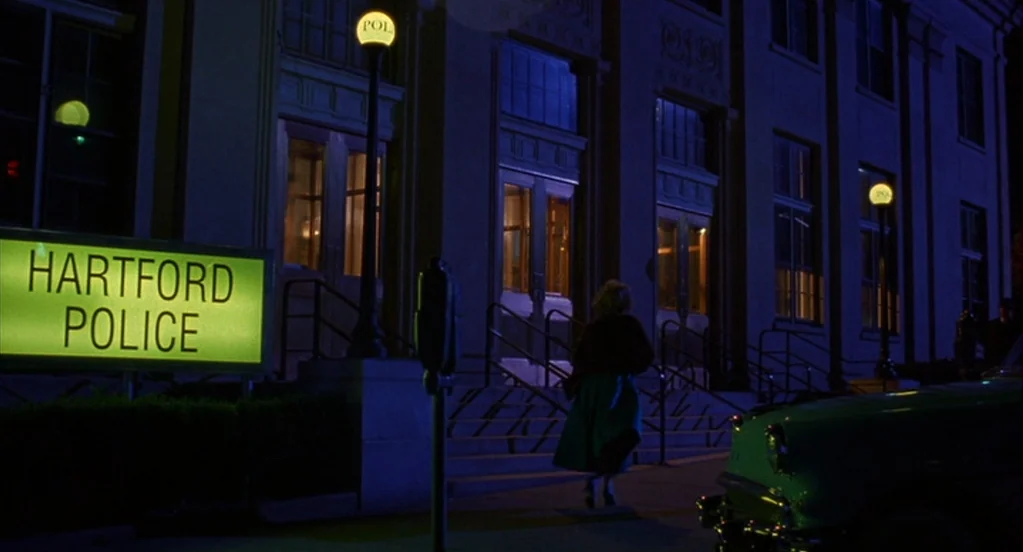 Far From Heaven (2002) movie still: wide — A dark blue night exterior features the stone facade of a local building. A woman walks toward the…