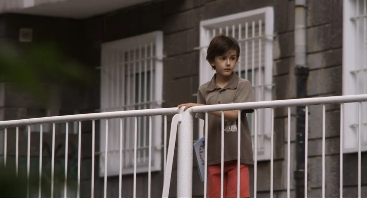 Newton's Third Law (2017) movie still: medium — A boy stands on a balcony in front of a stone building facade. He leans against a white metal…
