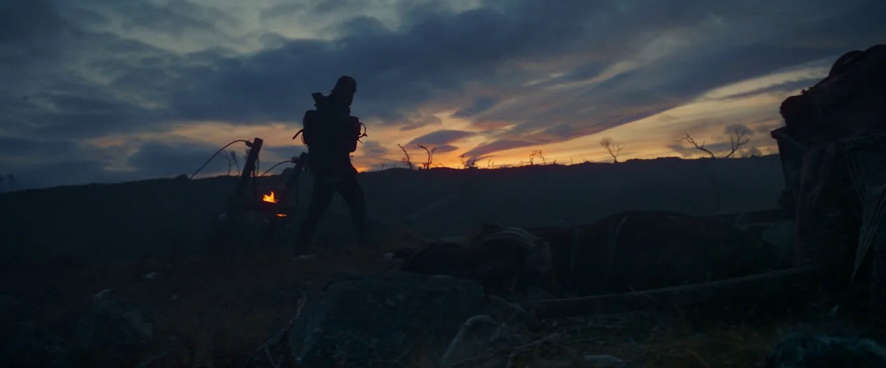 Sisu (2022) movie still: wide — A desolate and barren landscape extends beneath a dark, cloudy twilight sky. A solitary traveler moves across the…