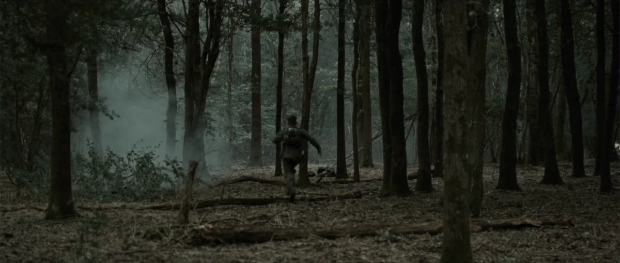 Sin retorno (2009) movie still: wide — A dense forest filled with tall thin trees and scattered fallen logs on ground. A man in tactical gear runs…