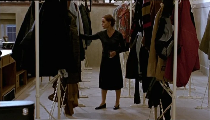 The Piano Teacher (2001) movie still: fullbody — A utilitarian cloakroom contains multiple free-standing garment racks filled with diverse winter…
