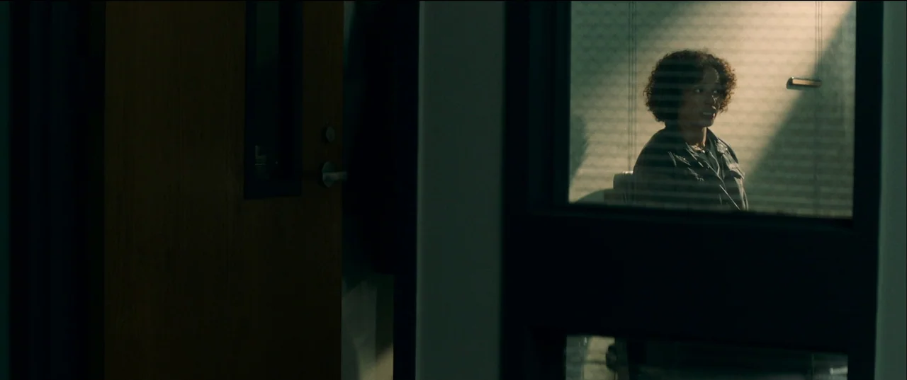 Birth/Rebirth (2023) movie still: medium — A dimly lit room with a door frame obscuring part of the view. A woman with curly hair sits in a chair…
