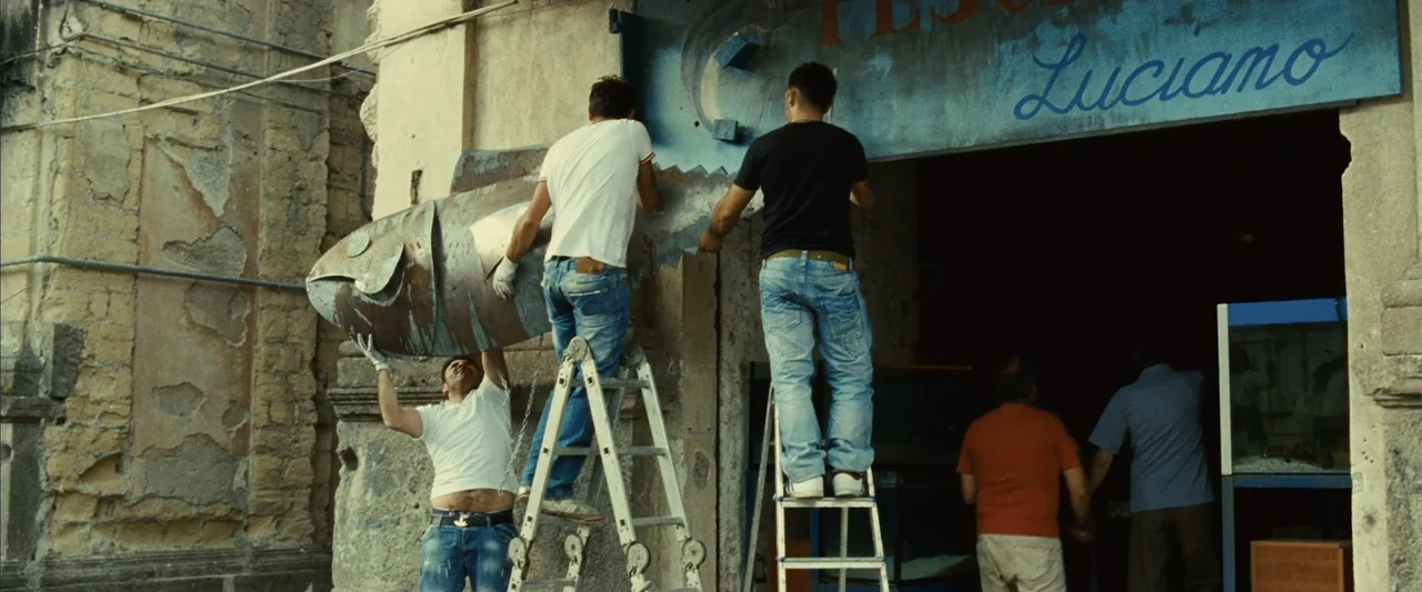 Reality (2023) movie still: medium — A weathered stone facade displays a blue sign for a shop named Luciano. Three men work on metal ladders to…