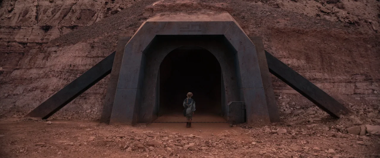 Settlers (2021) movie still: wide — An arid, rocky landscape features a large, industrial metallic entrance built into a cliff. A lone child stands…