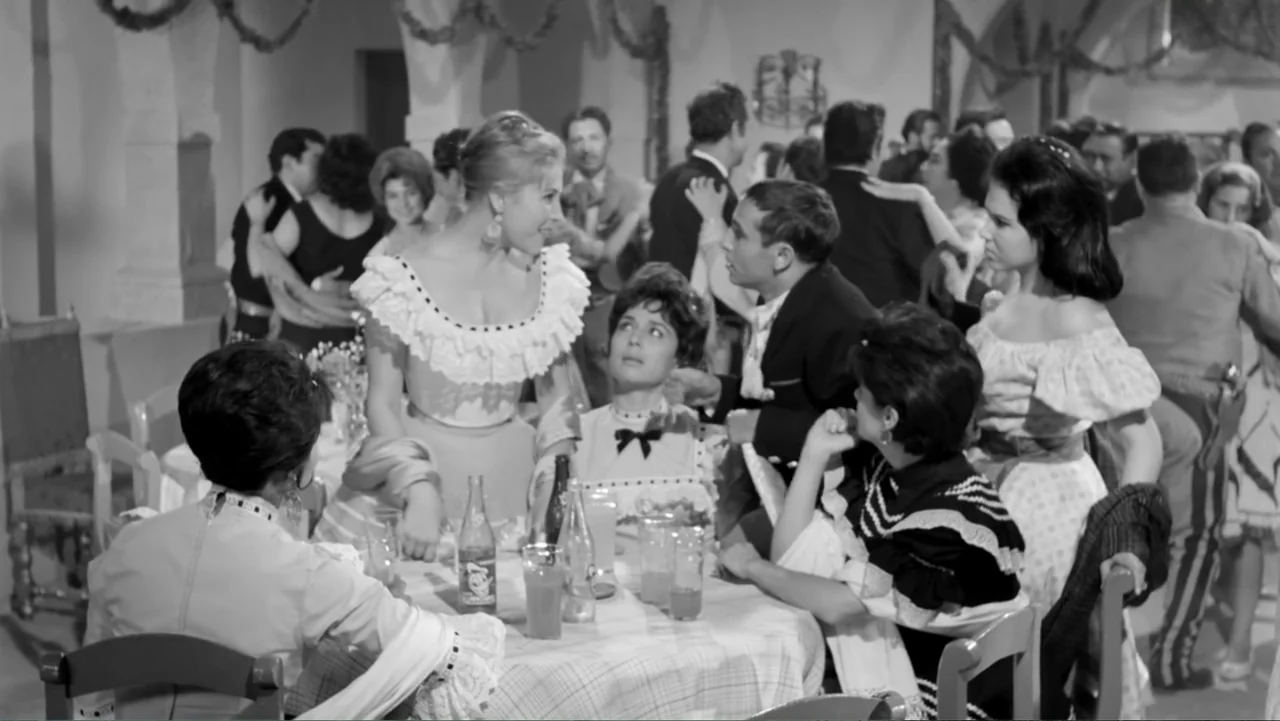 Escuela para solteras (1965) movie still: medium — A crowded hall features multiple tables with guests dressed in formal mid-century attire. A small…