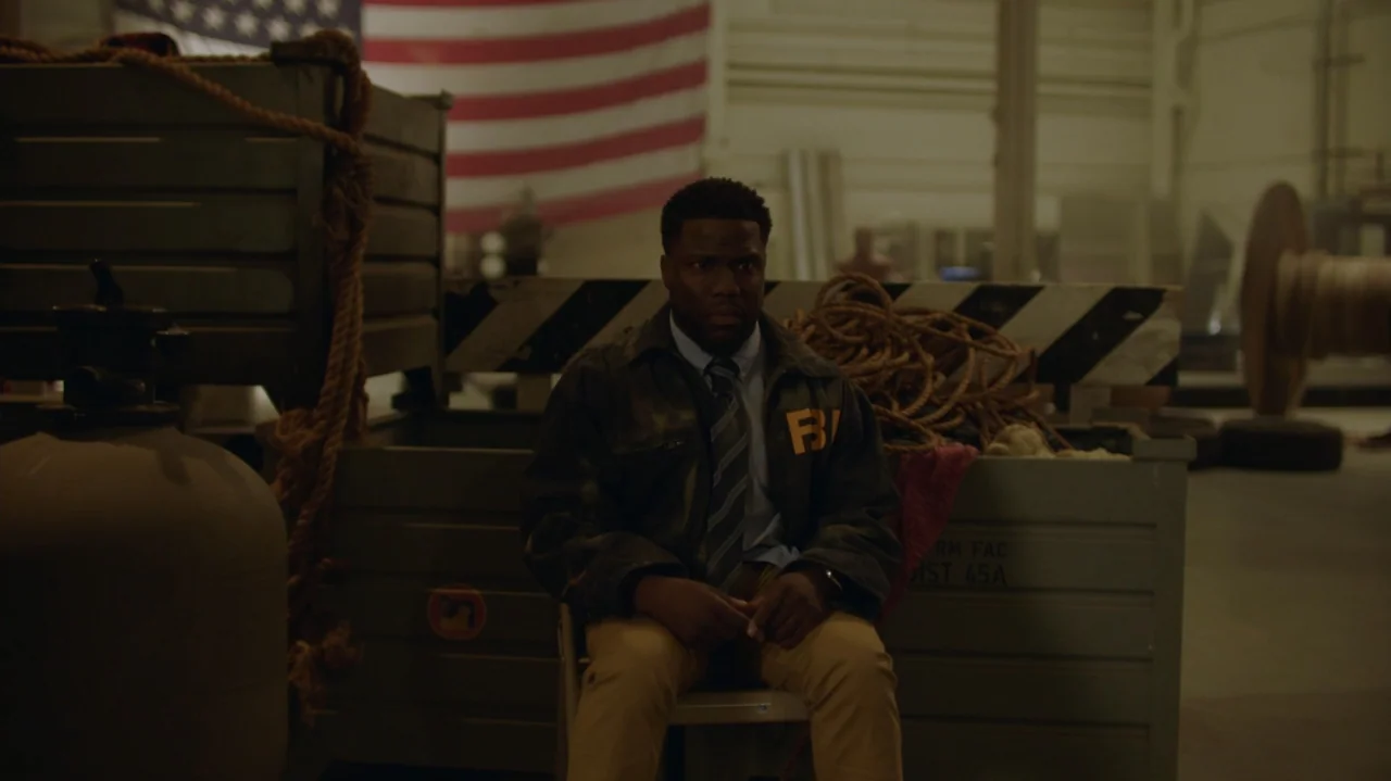 Die Hart (2023) movie still: medium — A dark industrial hangar contains large storage crates and an American flag. A man in an FBI jacket sits alone…