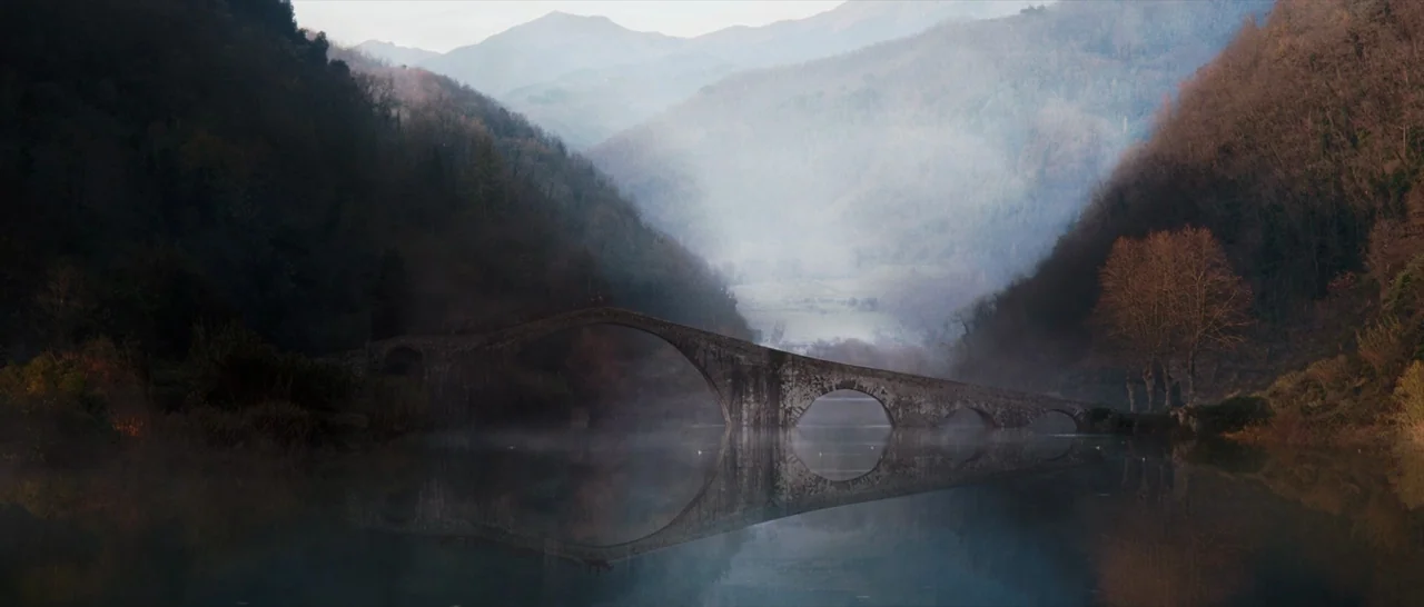 Tale of Tales (2015) movie still: establishing — An ancient stone bridge arches across a calm river between tree-covered mountain slopes. Thick…