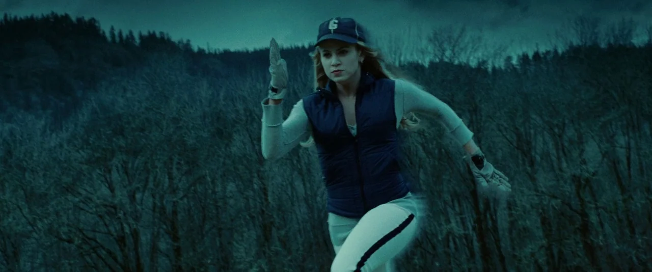 Twilight (2008) movie still: medium — A dense, dark forest of bare trees stretches into the distance under overcast skies. A blonde woman in…