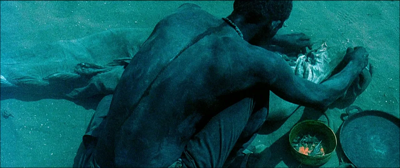 Black Hawk Down (2001) movie still: medium — A man crouches on dark sand near the water, working over a shrouded form. He tends to a small metal…