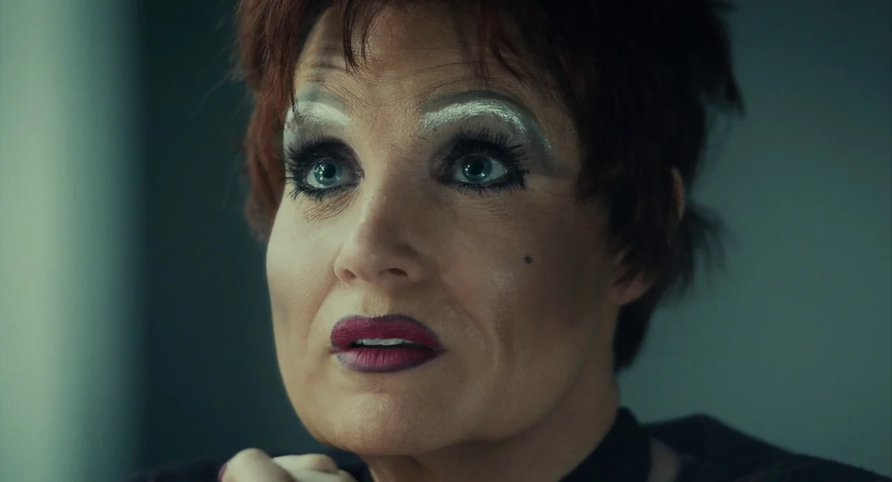 The Eyes of Tammy Faye (2021) movie still: closeup — A woman with stylized makeup sits in a dimly lit, cool-toned indoor space. She gazes upwards…
