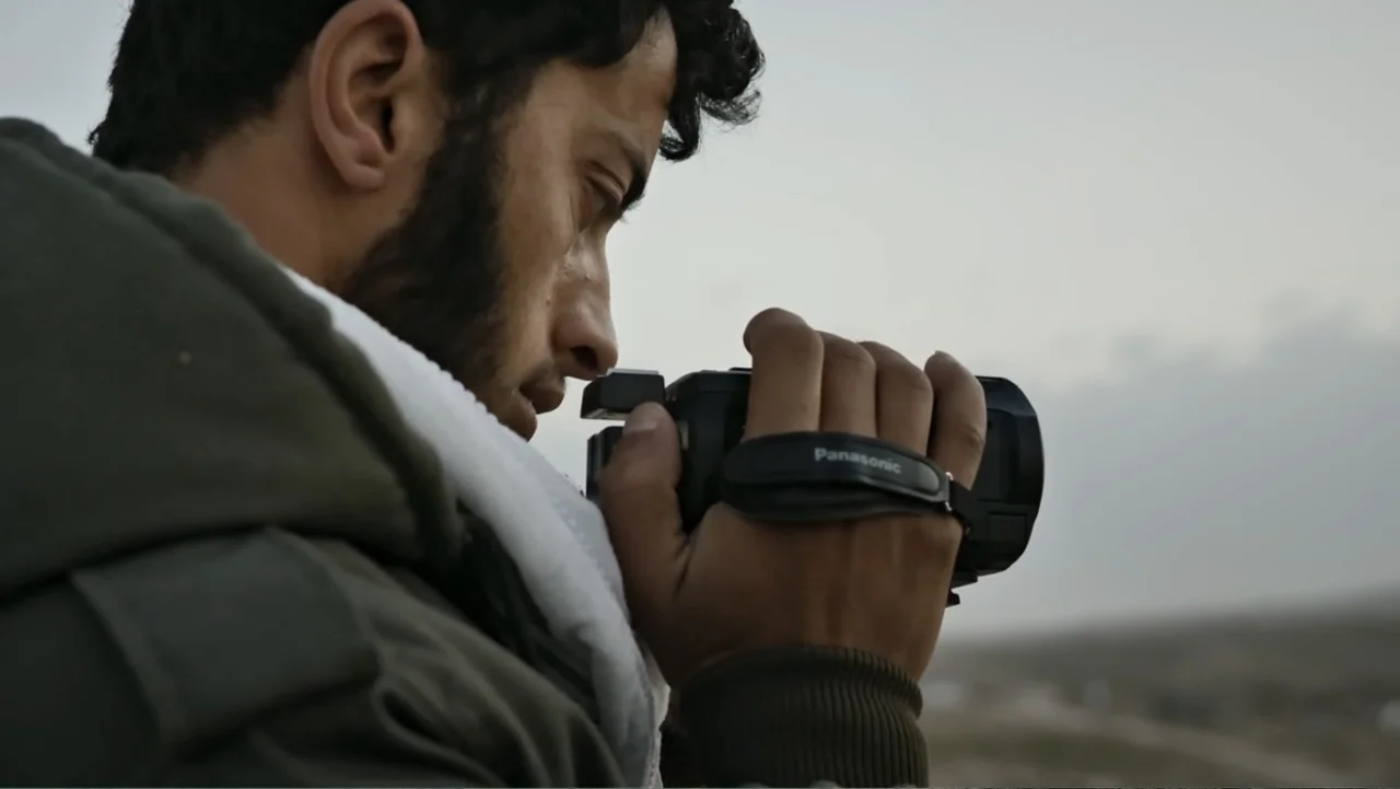 No Other Land (2024) movie still: closeup — A man stands outdoors in a desert landscape under an overcast sky. He holds a black handheld camcorder…