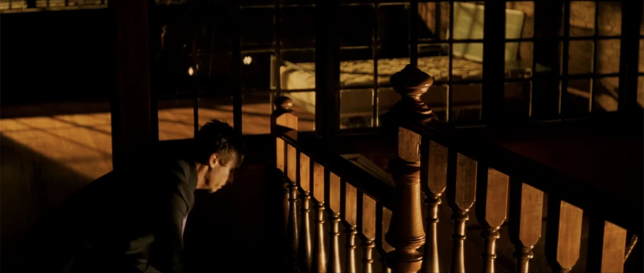 Elektra (2005) movie still: medium — A man stands near a wooden staircase landing inside a dimly lit building interior. The man looks down over the…