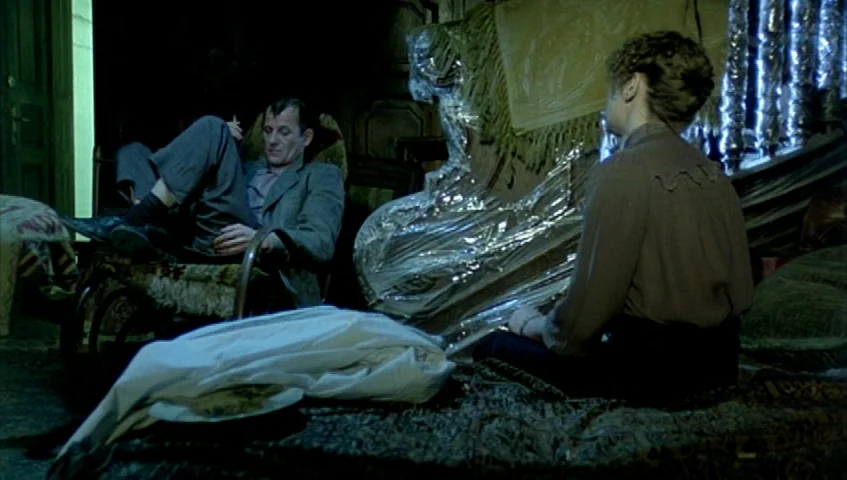 Almanac of Fall (1984) movie still: medium — A dimly lit room contains furniture covered in reflective plastic sheeting. A man reclines in a chair…