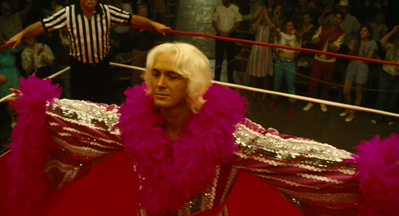 The Iron Claw (2023) movie still: medium — A crowded venue features a wrestling match with spectators gathered behind the ropes. A blonde man stands…