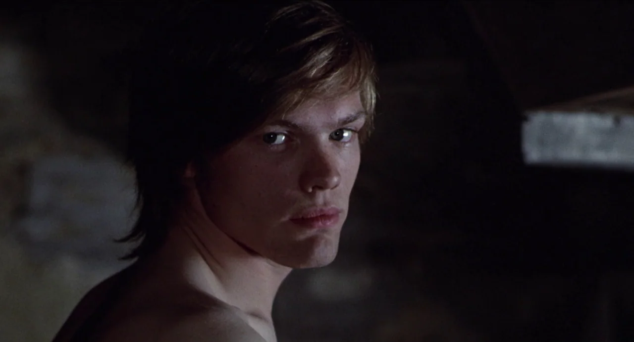 The Canterbury Tales (1972) movie still: closeup — A dimly lit interior space with a dark, indistinct background. A young man with light brown hair…