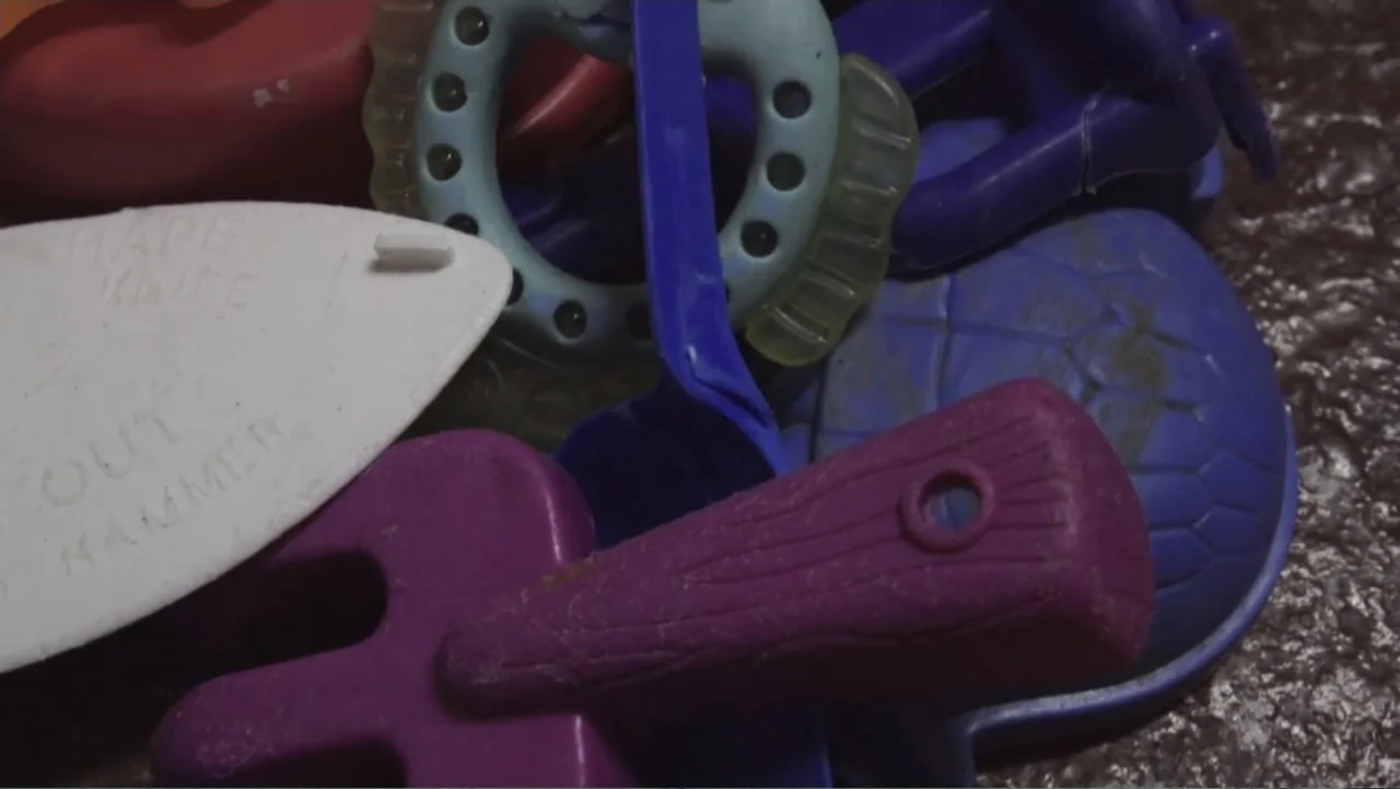 Hondar 2050 (2018) movie still: closeup — A jumble of colorful plastic toys sits on a dark textured surface. The items include shaped sand molds and…