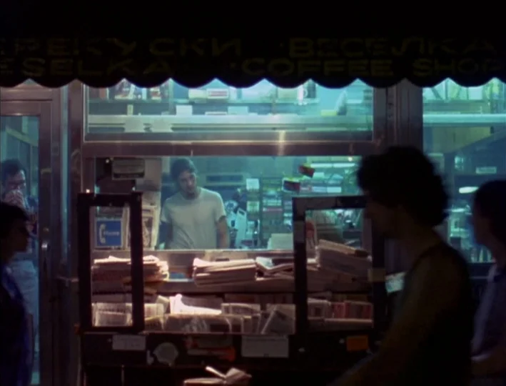 News From Home (1977) movie still: medium — A night newsstand interior is lit by overhead fluorescent lights casting a greenish tint. A man in a…