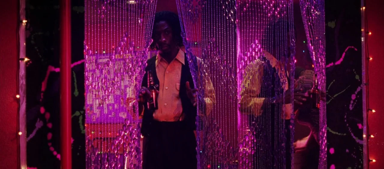 Showgirls (1995) movie still: medium — A man stands behind a thick magenta beaded curtain that obscures much of the view. He holds a brown glass…