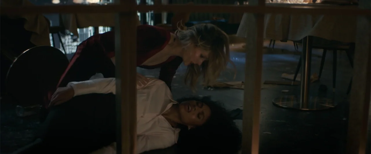 Drop (2025) movie still: medium — A dark restaurant floor area with furniture and debris scattered on the ground. One woman leans over another woman…