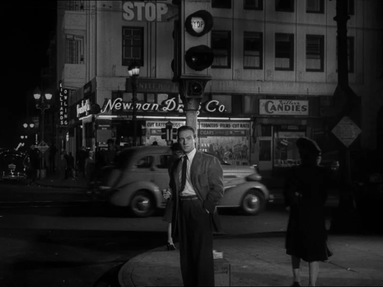 Double Indemnity (1944) movie still: fullbody — A night time city street corner features a drugstore with glowing window signage. A man in a suit…