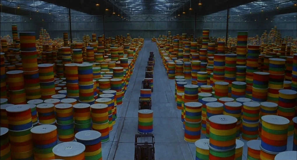 The Hudsucker Proxy (1994) movie still: wide — A vast industrial warehouse holds thousands of cylindrical containers stacked in tall, uniform…