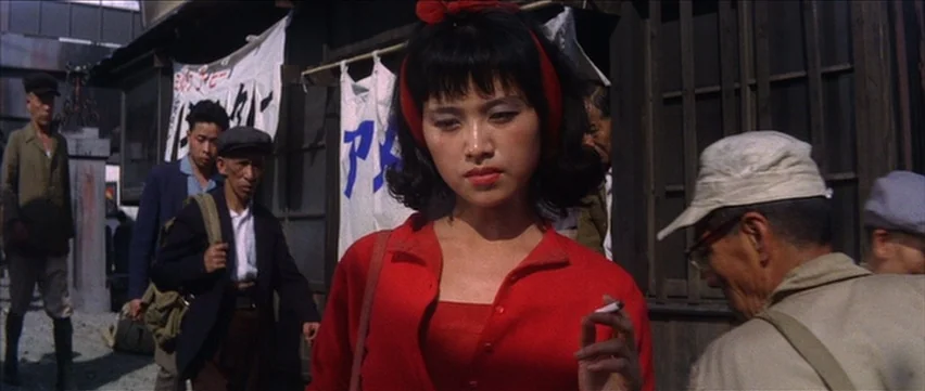 Gate of Flesh (1964) movie still: medium — A crowded outdoor space is lined with wooden structures and white banners with text. A woman in a red…
