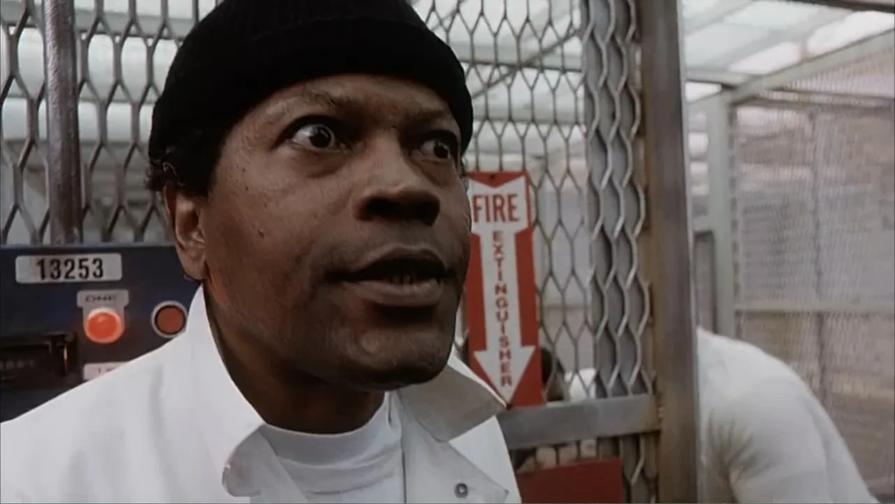 Against the Wall (1994) movie still: closeup — A prison facility featuring steel mesh fencing and red fire safety signage. A man in a knit cap…