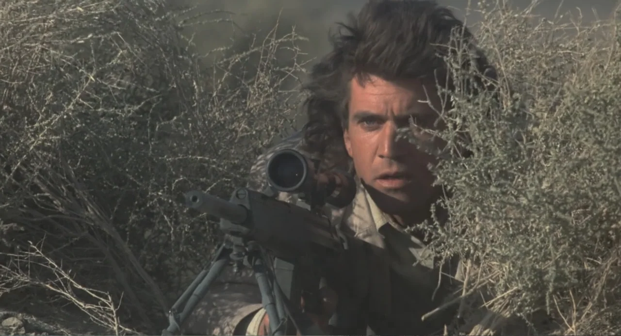 Lethal Weapon (1987) movie still: medium — A man hides among dry desert brush in an arid, open landscape. He is aiming a sniper rifle with a bipod…