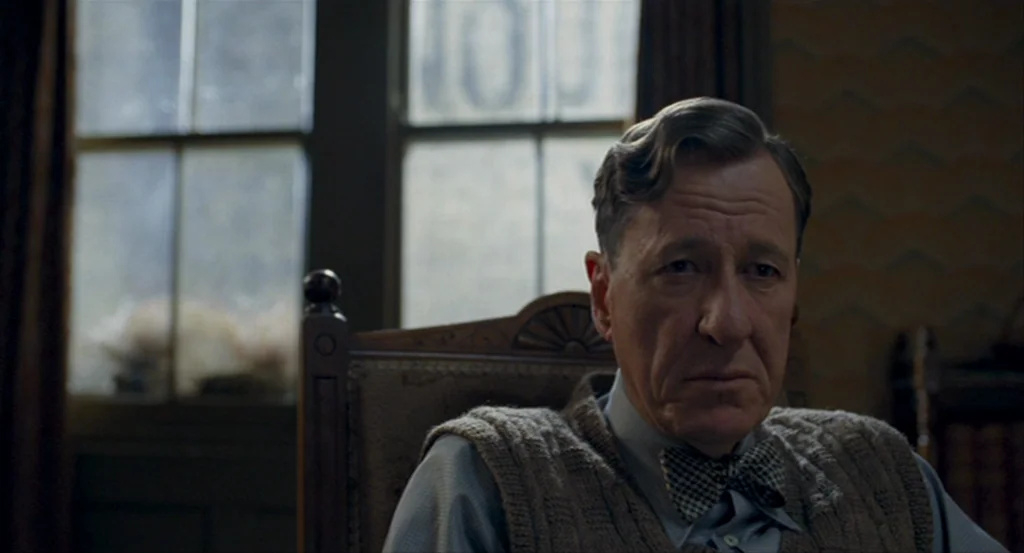 The King’s Speech (2010) movie still: medium — A man sits in a wooden chair near a window with glass panes. He is wearing a formal sweater vest and…