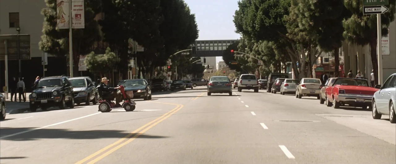 License to Wed (2007) movie still: wide — An urban intersection features tall trees lining the road and numerous parked automobiles. An older man…