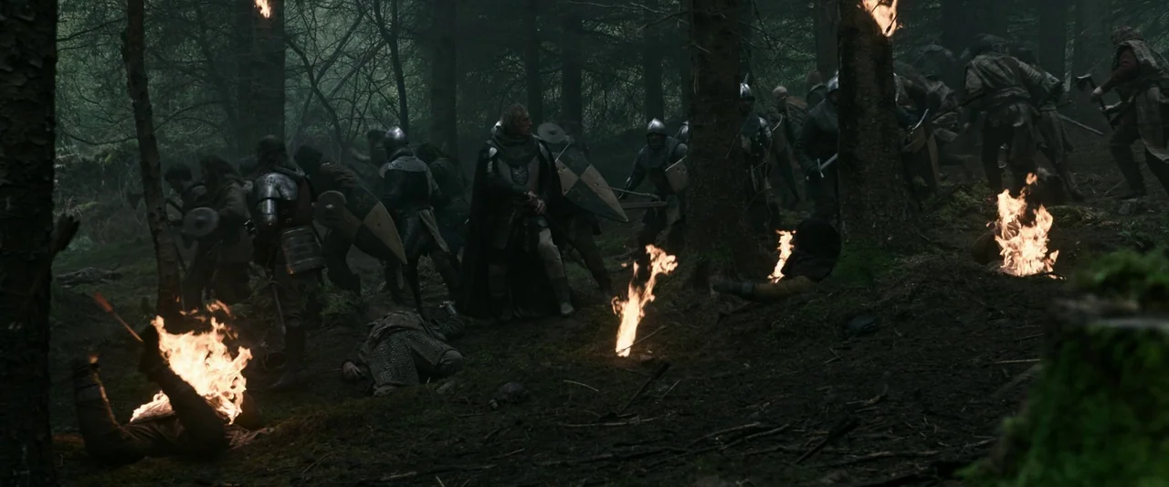 The Last Duel (2021) movie still: wide — A dark, dense forest contains several fires burning on the ground and against tree trunks. Multiple…