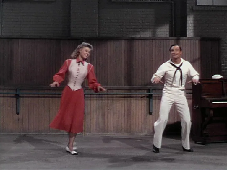 On The Town (1949) movie still: fullbody — A dance studio interior features wooden wall paneling and a dark upright piano nearby. A woman in a red…