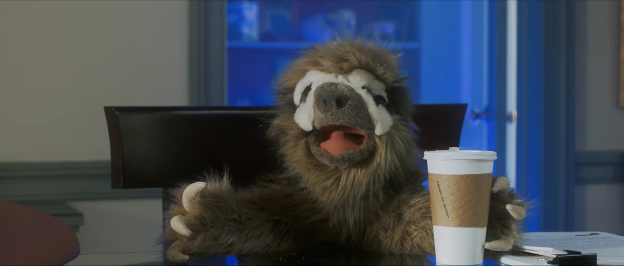 Songs For A Sloth (2021) movie still: medium — A furry sloth puppet sits at a glass desk in a dark room. The puppet holds a paper coffee cup with a…