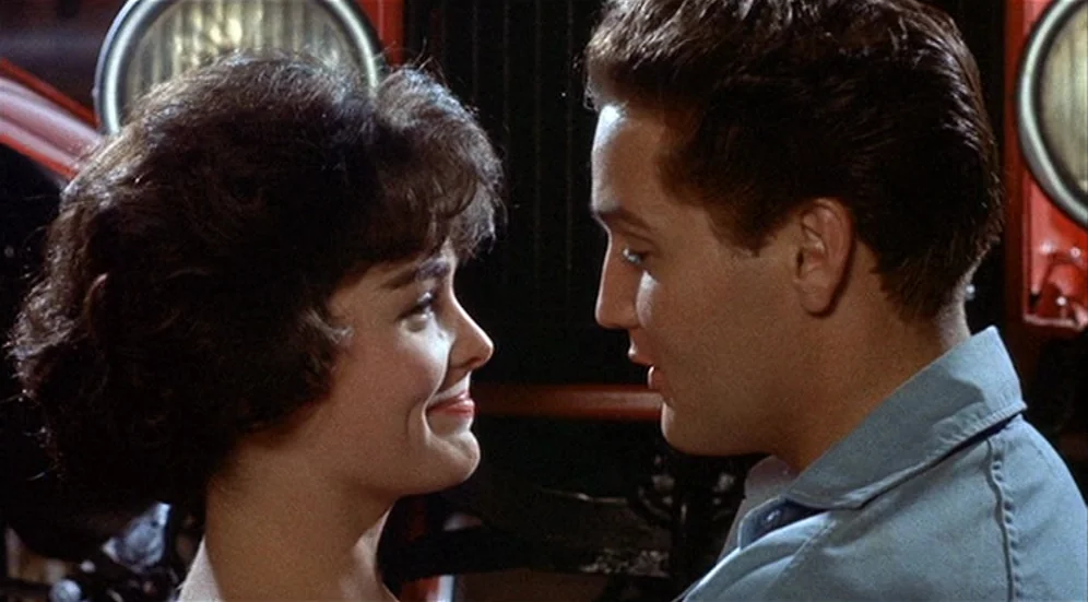 Kid Galahad (1962) movie still: closeup — Two young people face each other in front of a red vintage vehicle. The man and woman exchange eye contact…
