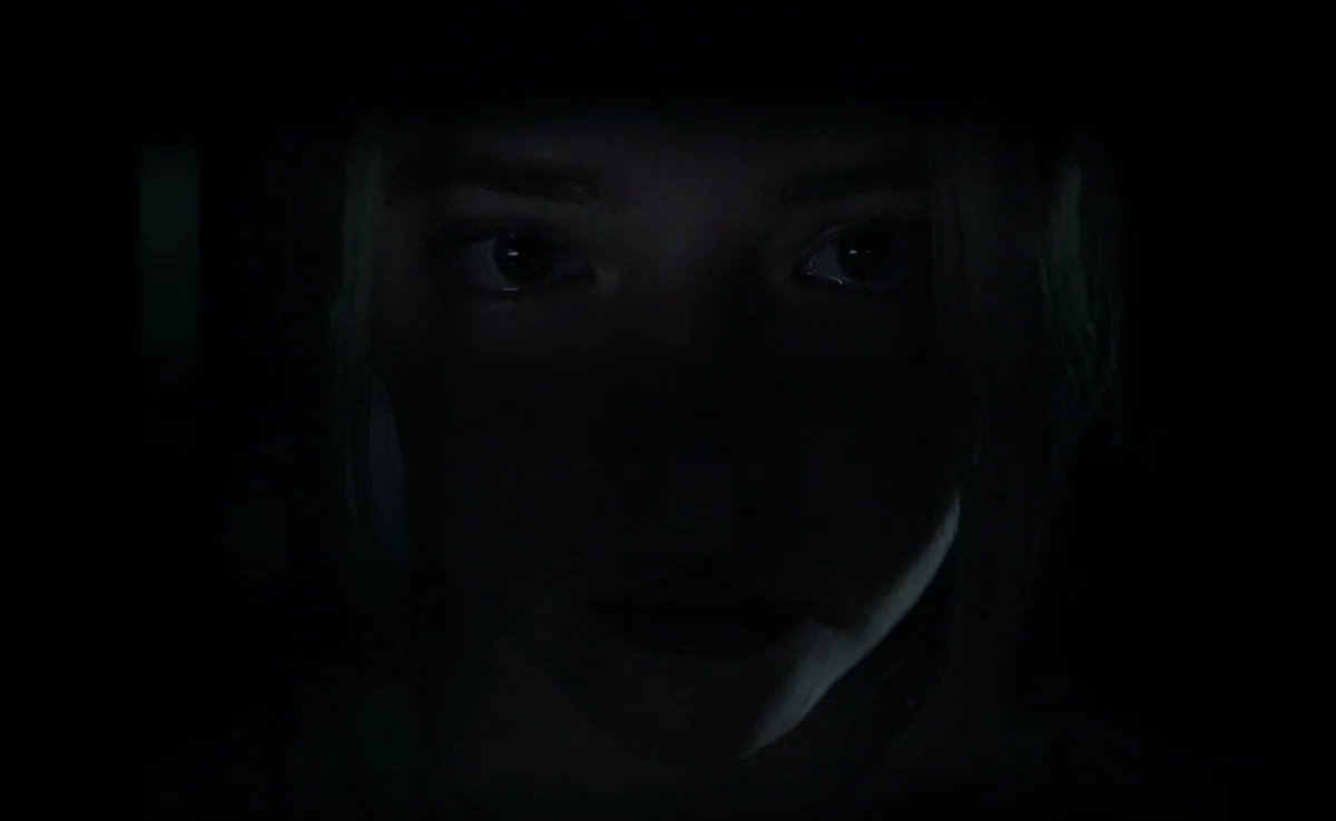 The Witch (2015) movie still: closeup — The face of a young woman is partially lit in an extremely dark environment. She stares forward while her…