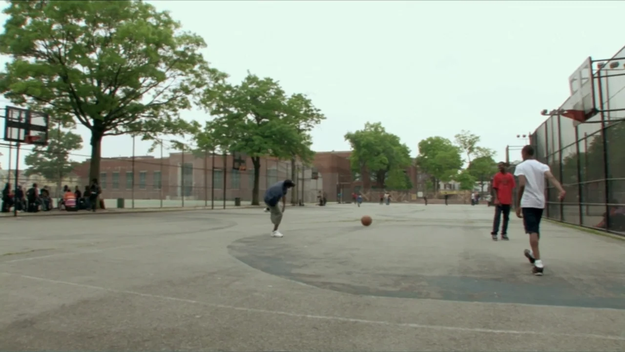 Little White Lie (2008) movie still: wide — A fenced urban basketball court is surrounded by large trees and brick school buildings. A young man…