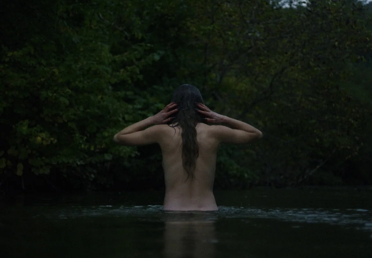 You Won’t Be Alone (2022) movie still: medium — A dark river flows against a backdrop of dense, shadowy foliage at dusk. A woman stands waist-deep…