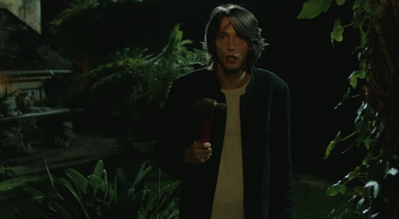 Cosmos (2015) movie still: medium — A dark, vegetation-filled outdoor space at night with large leaves in the foreground. A young man stands looking…