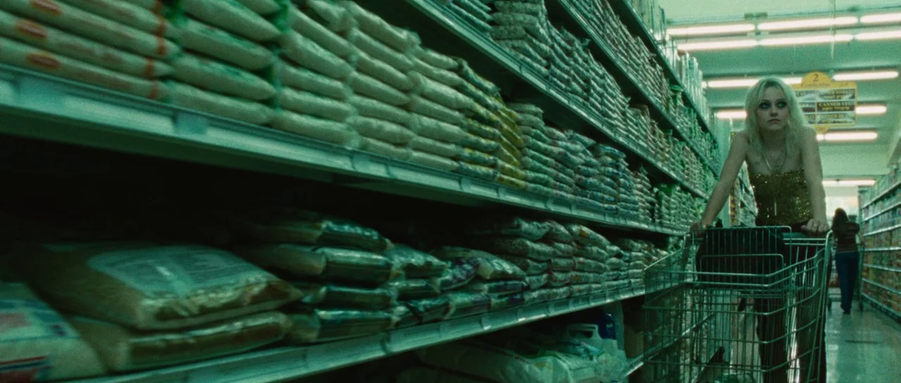 The Runaways (2010) movie still: medium — A long grocery store aisle is filled with stacked bags of white rice. A young woman with blonde hair…
