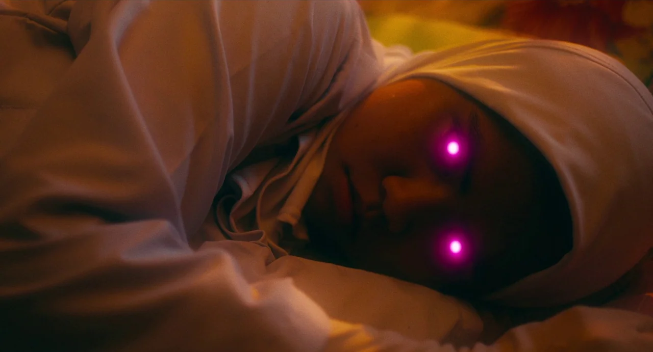 Tiger Stripes (2023) movie still: closeup — A person lies on a soft surface in a darkened environment. Glowing magenta light emits directly from…