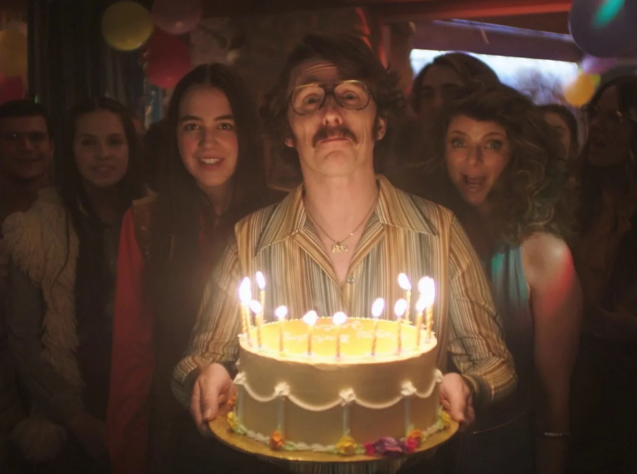 Girl Asleep (2015) movie still: medium — A dimly lit indoor party environment with colorful balloons hanging from the ceiling. A man holds a lit…