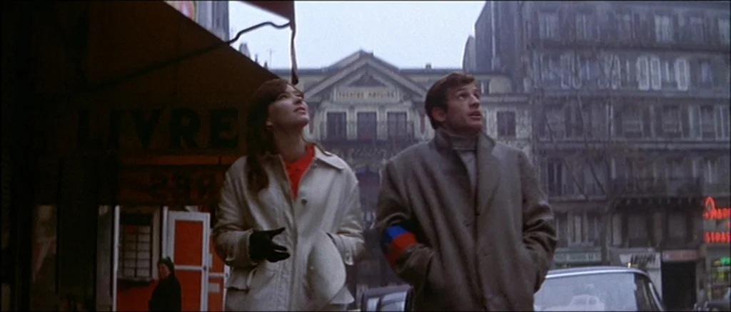 Une Femme Est Une Femme (A Woman Is a Woman) (1961) movie still: medium — An urban street scene featuring a woman and man looking at a building. An…
