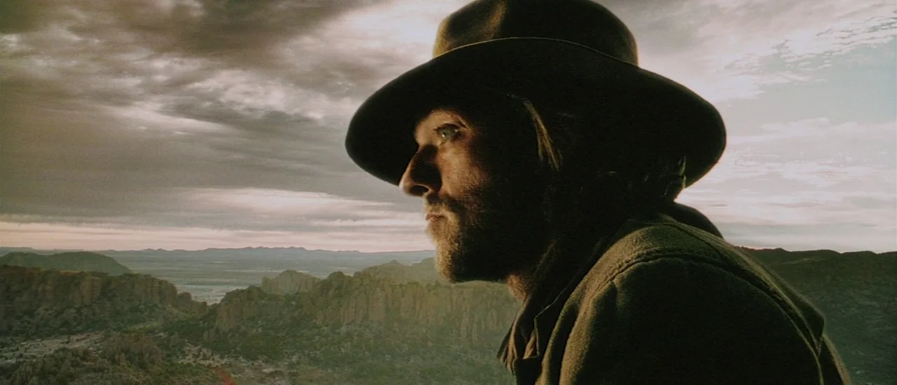Renegade (2004) movie still: closeup — A vast, rugged mountainous desert landscape stretches beneath a cloudy golden hour sky. A man wearing a hat…