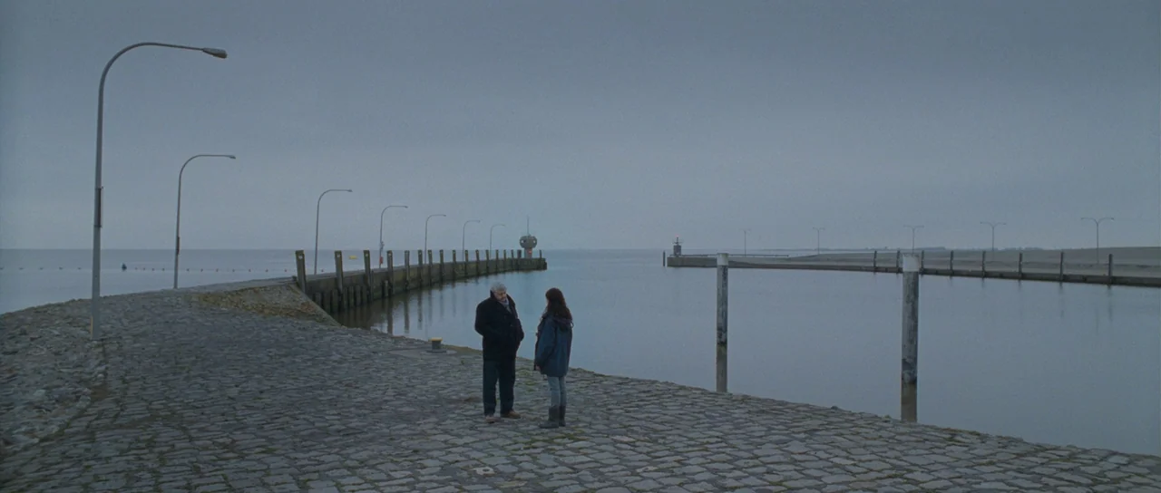 Womb (2010) movie still: wide — A long paved stone jetty extends into a calm, gray body of water. An elderly man and a woman stand together on the…